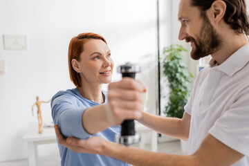 happy woman looking at physiotherapist while training arm with dumbbell in rehab center.