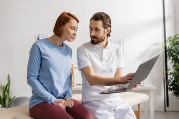 bearded physiotherapist with laptop talking to smiling woman during appointment in rehabilitation center.