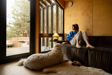 Woman sitting relaxed on a couch by the window with her dog at wooden cottage in forest. Concept of...