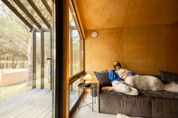 Woman cares her dog while sitting on a couch by the window, spending leisure time together in...