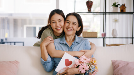 Happy time Mother day grown up child looking at camera cuddle hug give flower gift box red heart card to mature mum. Love kiss care mom asia middle age adult people smile enjoy sitting at home sofa.