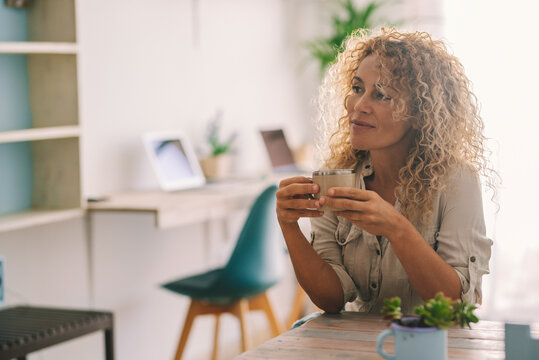 Attractive woman with long blonde curly hair enjoy time in indoor home leisure activity alone drinking a coffee or tea and looking away dreaming and thinking. Serene female people in relax time inside