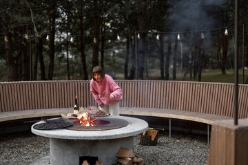 Young woman prepares food on beautiful outdoor bbq area with fire and round bench in pine forest at dusk. Luxury lifestyle at countryside concept