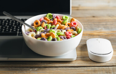 Colored breakfast cereals in a bowl and laptop, workplace breakfast, quick healthy breakfast in the office in the morning.