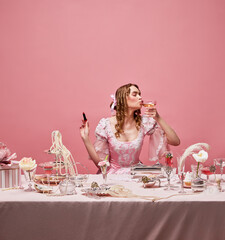 Shot of adorable blond princess wearing fancy pink dress and sitting at beautifully laid table with wine glass and lipstick over studio background. Drinking cocktail - obrazy, fototapety, plakaty