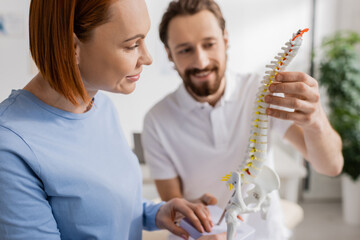 smiling chiropractor showing spine model to redhead woman during appointment in consulting room.