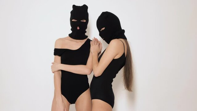 Two beautiful sexy women in black swimwear bathing suit. Models wearing bandit balaclava mask. Hot girls posing near white wall in studio. Seductive female in nice lingerie. Crime and violence