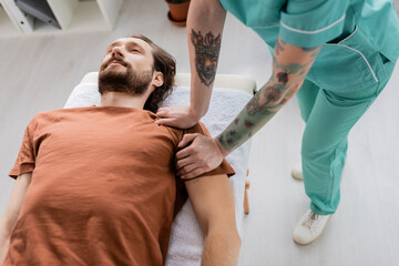 tattooed chiropractor doing shoulder massage to injured bearded man lying in consulting room.