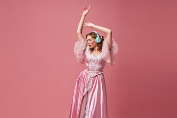 Naklejka premium Shot of adorable blond queen wearing headphones and dress listening music over pink studio background. Modern princess