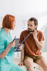 Obraz premium redhead physiotherapist with clipboard talking to bearded man touching injured neck in consulting room.
