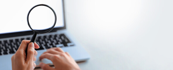Hand holding magnifying glass in front of a laptop screen. Symbol of web browsing or zooming displeyed something on screen. Paying attention on detail.