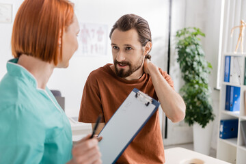 bearded man touching painful neck near blurred physiotherapist with clipboard in consulting room.