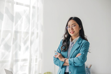 Portrait of young Asian woman in casual clothes standing with arms crossed, smiling and looking at camera while standing in modern office.