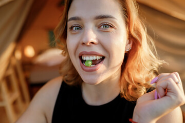Smiling Woman freelancer eating grape in cozy glamping tent in a sunny day. Luxury camping tent for...