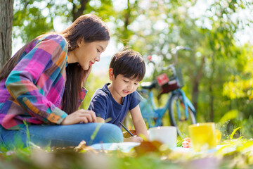 Happy cheerful Asian lovely little boy and mother sitting under the tree at park in the morning and drawing a picture. Asian woman spending time in weekend with her lovely little son.