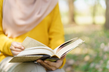 Happy cheerful Asian beautiful Muslim woman reading a book - Koran under the tree. Female muslim student studying an Islam Koran.