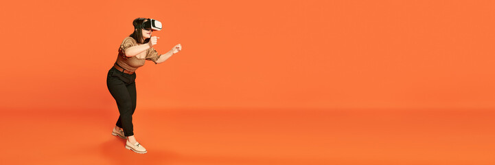 Portrait of young girl wearing VR glases and playin g online vide games against orange studio background. Driving. Concept of emotions, lifestyle, youth, modern technologies. Banner. Copy space for ad