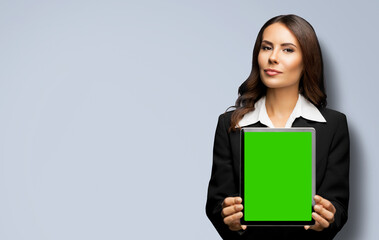 Obraz premium Portrait of young brunette businesswoman showing tablet pc, touchpad, with green chroma key screen, on grey gray background. Confident attractive business woman at office.