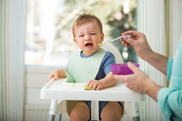 Father feeding his cute baby with first solid food, infant sitting in high chair. Child doesn't want to eat, refuses eating, making faces. Cozy kitchen interior. Healthy food concept. 