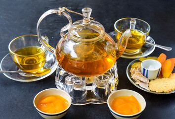 Glass tea pot with plum mango tea and ham,cups and crystalized fruits on black background
