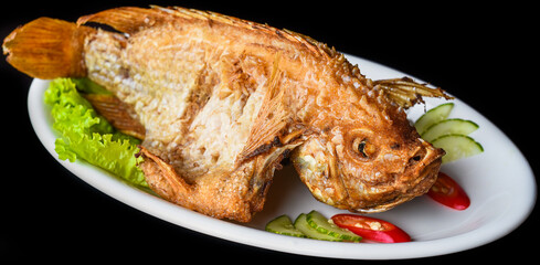 Fish grilled in batter with vegetables and greens on white plate isolated on black background side view