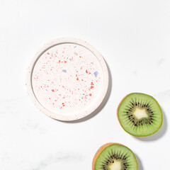 Fototapeta na wymiar Beauty cosmetics product presentation flat lay mockup scene with beige terrazzo circle shape and kiwi fruits on white marble table with copy space. Trendy sunlight, square format. Studio photography.