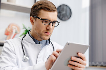 Male Caucasian doctor in white medical uniform filling patient medical history or anamnesis on tablet in office. Doctor consulting client online on tablet in modern clinic, healthcare concept - obrazy, fototapety, plakaty