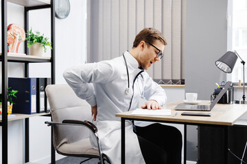 Fototapeta na wymiar A young male doctor sits at his workplace and feels pain in his back. A doctor takes a break from his work day because of pain