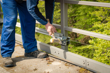 A worker around 60 years old hits a fastener for a cable duct with a non rebound soft face hammer. A cable duct is installed on a railing high up in the height of treetops.