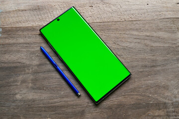 Smartphone mockup with green screen, stylus pen on the side, top view, isolated on wooden background