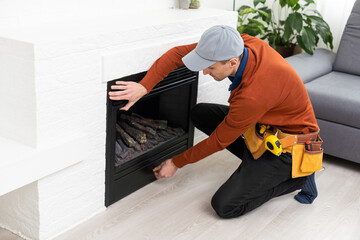 Professional technician sealing electric fireplace with caulk near white wall in room