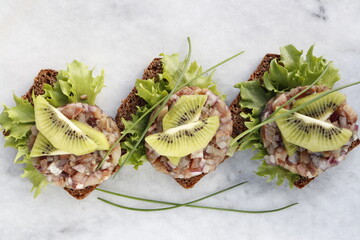 Three slices of rye bread with herring tartare, garnished with kiwi slices.