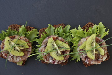 Three slices of rye bread with herring tartare, garnished with kiwi slices.