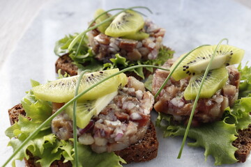 Three slices of rye bread with herring tartare, garnished with kiwi slices.