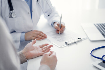 Fototapeta premium Doctor and patient sitting at the table in clinic. The focus is on female physician's hands filling up the medication history record form or checklist, close up. Medicine concept