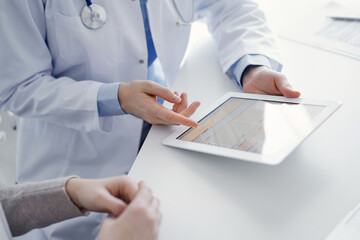 Doctor and patient sitting at the table in clinic. The focus is on female physician's hands using tablet computer, close up. Medicine and healthcare concept