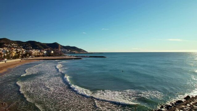 Panoramic drone aerial view of coastal city, old town of Sitges seen during sunny day. Close-up of surging Mediterranean sea waves. Province of Barcelona, Catalonia, Spain. Film intro. 4K video
