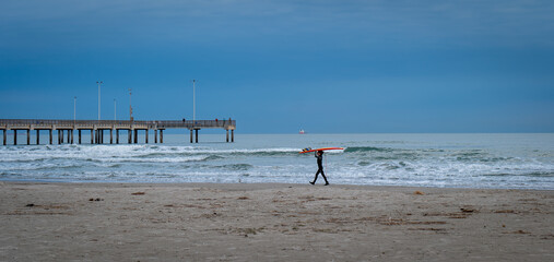 PORT ARANSAS, TX - 9 FEB 2023: Young surfer carries a surfboard on the beach, near the water of the Gulf of Mexico and a pier, in evening light.