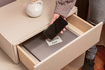 Close up of hand holding smartphone in separate desk drawer labeled Device Free Zone. Woman putting...