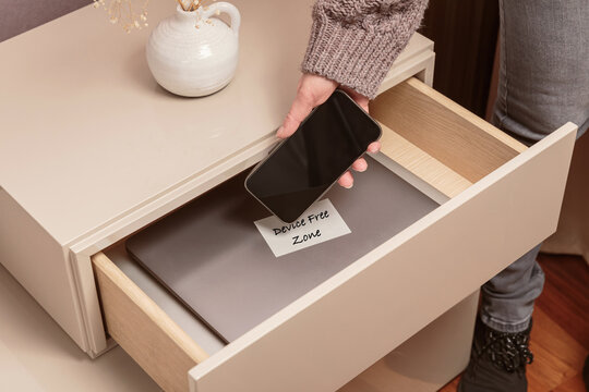 Close up of hand holding smartphone in separate desk drawer labeled Device Free Zone. Woman putting her phone in drawer with different gadgets at home. Digital detox and technology addiction concept