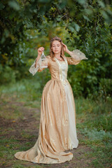 A beautiful young woman in a historical golden dress with a crown on her head holds a white cage in her hands. Princess in a medieval dress in the forest.