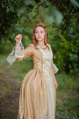 A beautiful young woman in a historical golden dress with a crown on her head holds a white cage in her hands. Princess in a medieval dress in the forest.