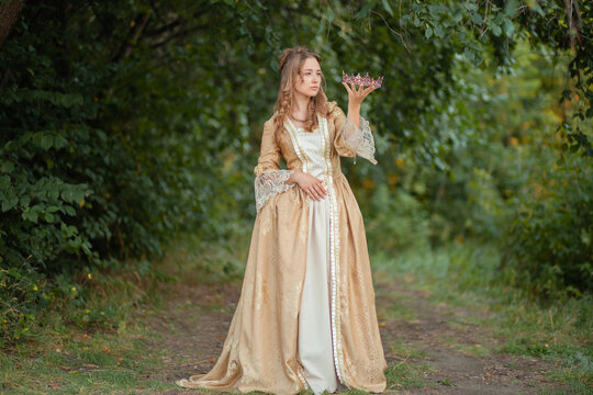 Beautiful young woman in a historical gold dress with a crown in her hands. Princess in a medieval dress in the forest. - Powered by Adobe
