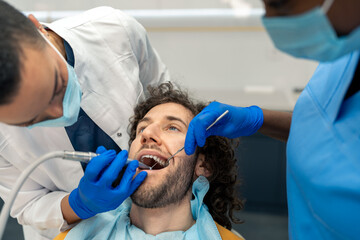 Female dentist specialist examining and working on patient's teeth with her dental assistant using dental drill, removing dental plaque, preparing teeth for dental filling at dentist's office.