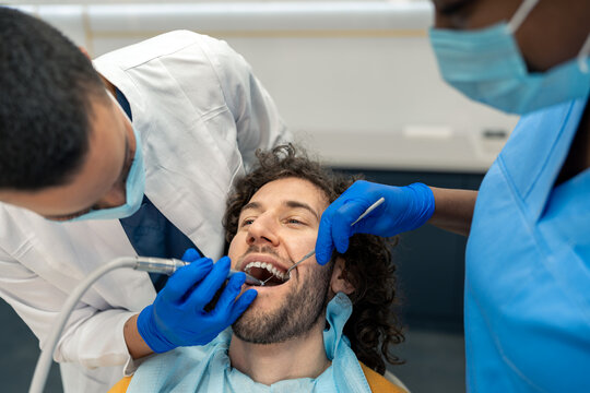 Young man having dental drill procedure at dentist's office. Female dentist specialist and dental assistant working on patient's teeth using dental drill, removing dental plaque.