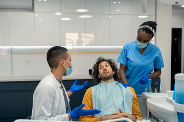 Anxious male patient on his first appointment with his dentist and her assistant dental nurse with surgical masks discussing future dental procedures and treatment for healthy gums and teeth.