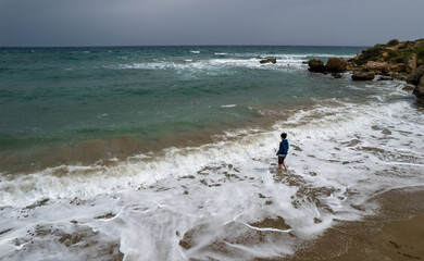 Drone aerial of  young boy  in  shorts standing on beach and looking at the waves. View from the back. Stormy weather on the sea coast. Stormy summer day.