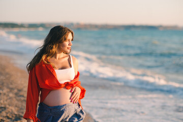 Fototapeta na wymiar Pregnant happy woman walking at sea shore over sun light wearing casual prenatal clothes at beach. Motherhood. Summer vacation season.