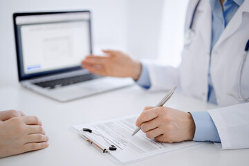 Doctor and patient sitting opposite each other at the desk in clinic. The focus is on female physician's hands filling up the medication history record form or checklist, close up. Medicine concept