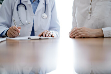 Doctor and patient sitting near each other at the wooden desk in clinic. Female physician's filling...
